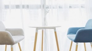 Een kamer met een witte tafel en twee stoelen, één wit en één blauw, met uitzicht door dunne gordijnen.