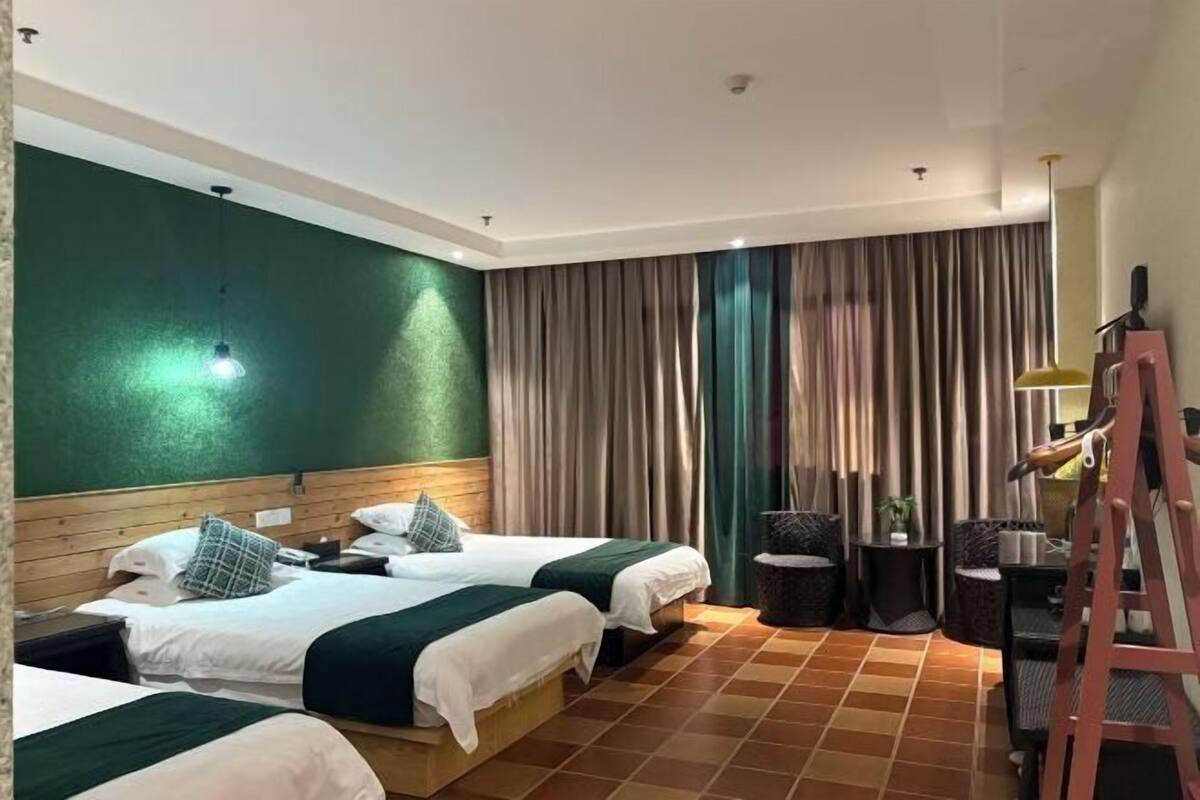 A hotel room with two beds, a wooden headboard, a green accent wall, a large window with curtains, a small table with chairs, and a ladder.