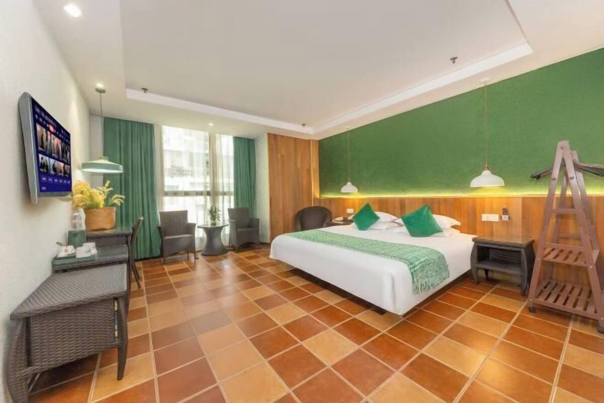 A hotel room with a large bed, green curtains, a desk with a chair, a small table, and a ladder.