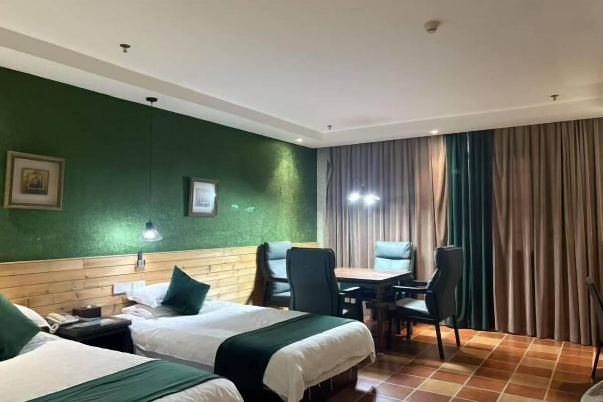 A hotel room with two beds, a wooden headboard, a table with chairs, a lamp, and a large window with curtains.