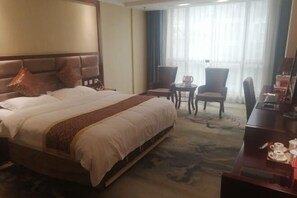A hotel room with a large bed, a desk with a chair, a small table, and a window with curtains.
