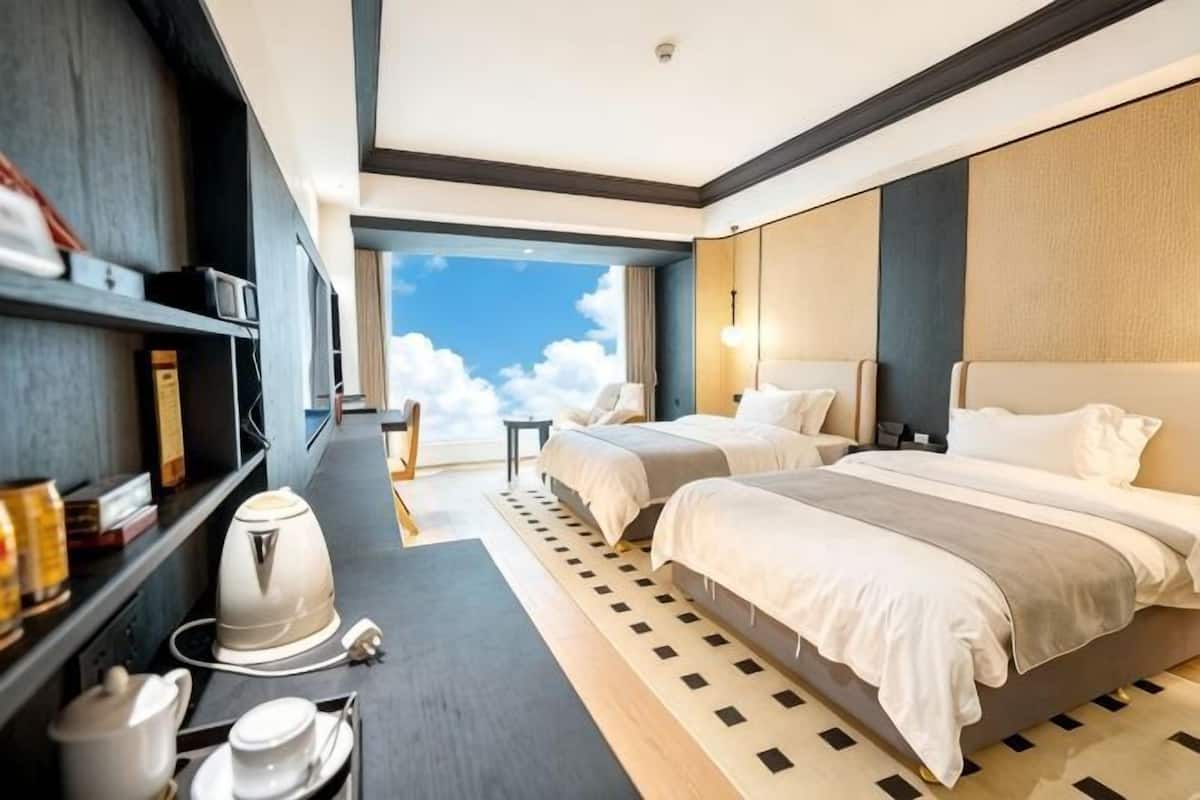 Habitación de hotel con dos camas, un escritorio con hervidor, una estantería y un ventanal con vistas al cielo.