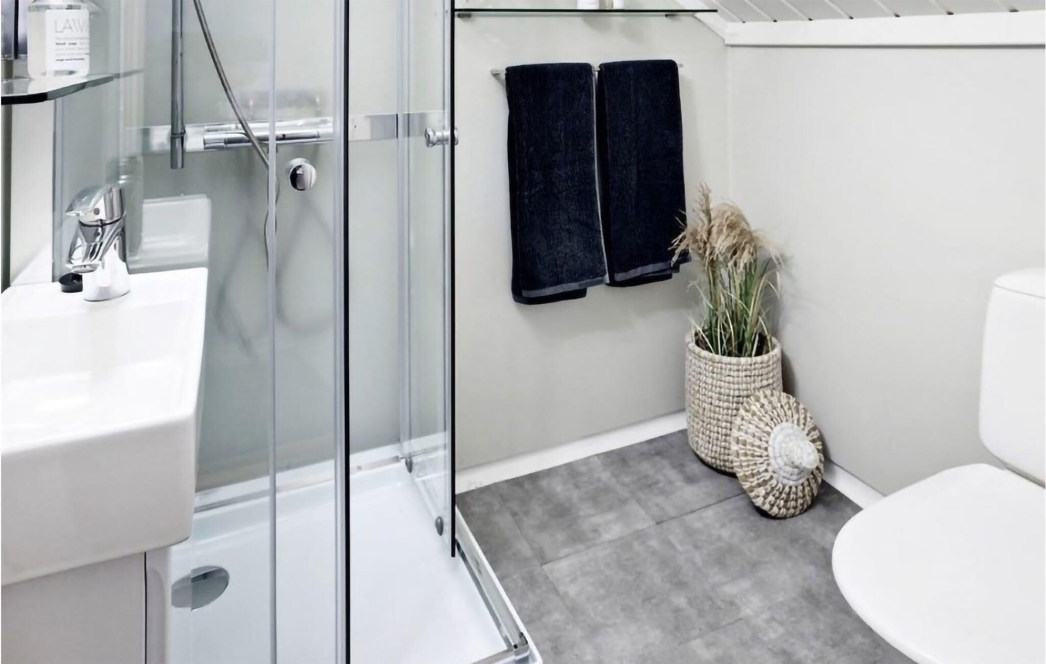 A modern bathroom with a glass shower enclosure, a white sink, and a toilet. There are two black towels hanging on the wall and a woven basket with dried plants next to the toilet.