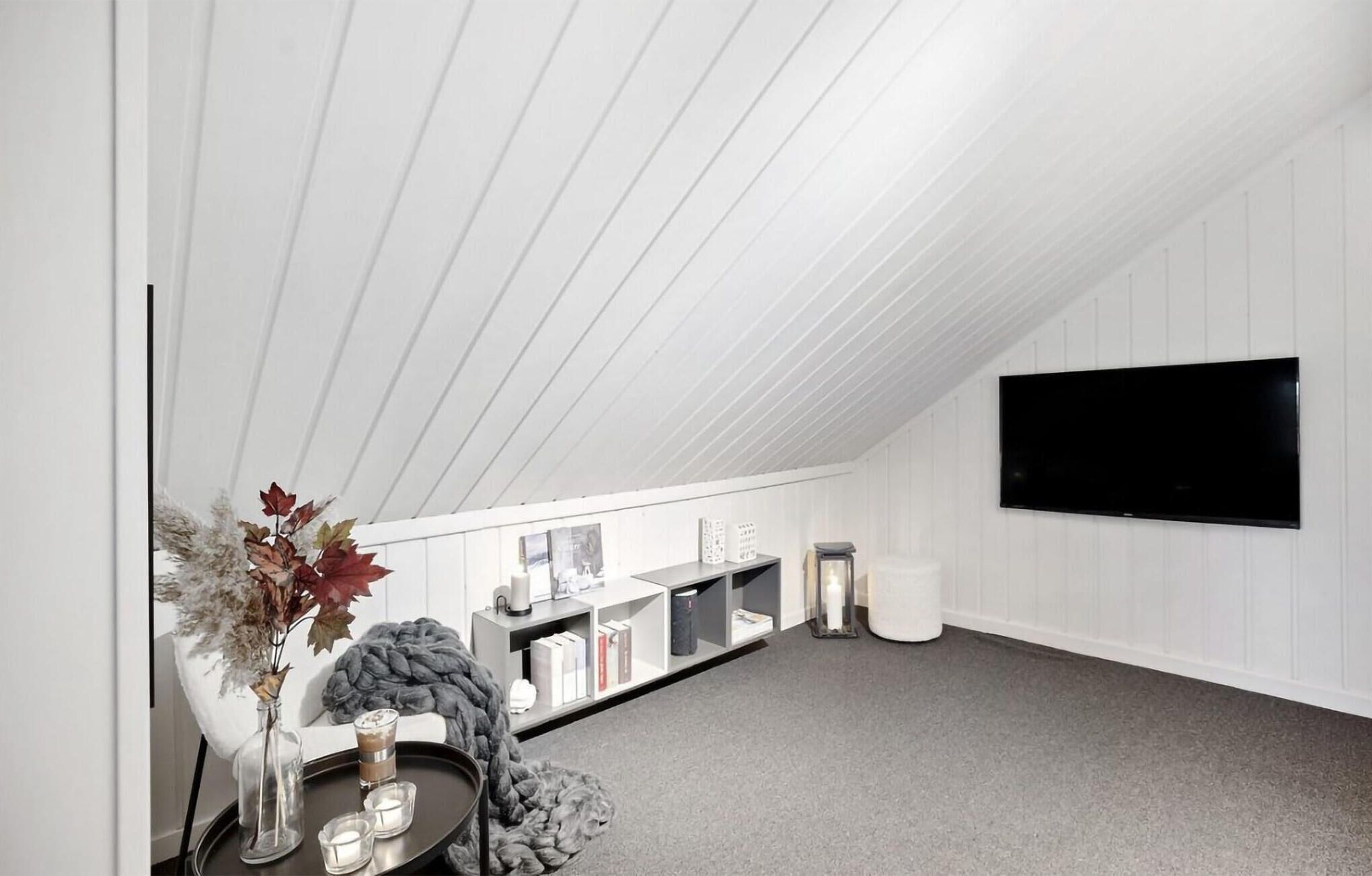 A modern loft bedroom with a sloped ceiling, a mounted TV, a bookshelf, and a cozy seating area with a blanket and candles.