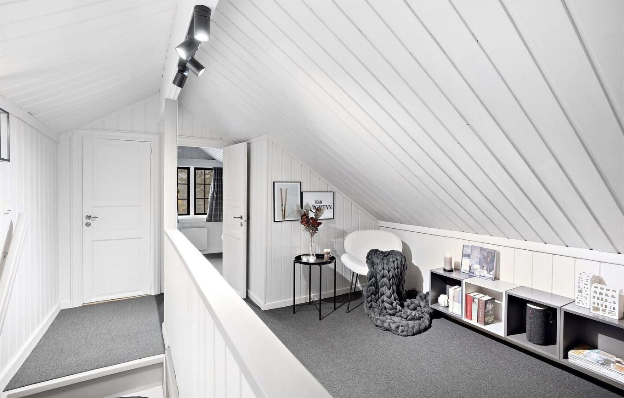 Een zolderkamer met witte wanden en een schuine kap, een witte stoel, een klein rond tafeltje met een vaas, en een boekenkast met boeken en decoratieve objecten.