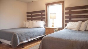 Una habitación con dos camas, cada una con cabecera y almohadas, una mesita de noche con lámpara y una ventana con persianas.