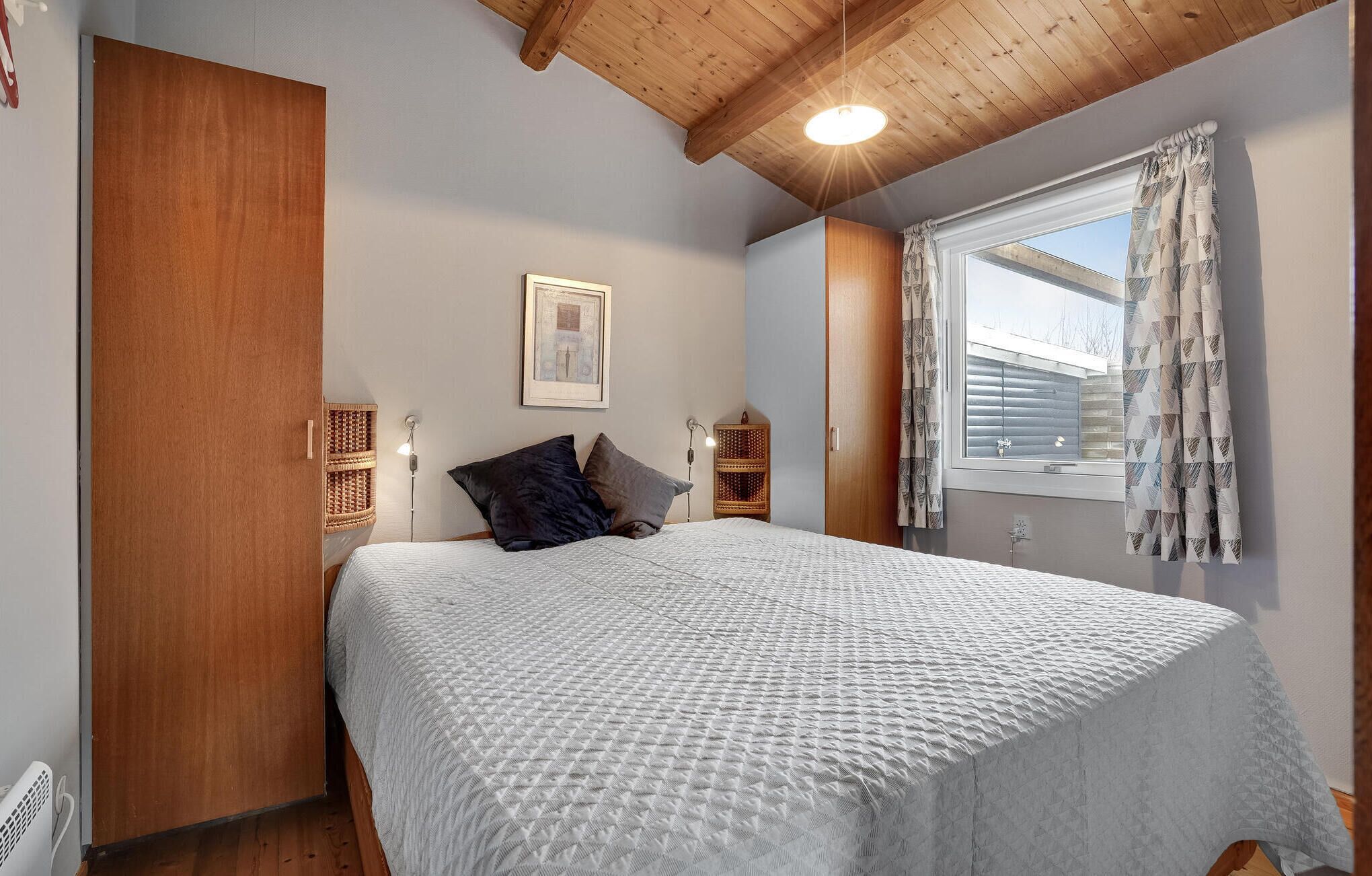 Una camera da letto con un letto, un armadio in legno, tavolini di vimini, un quadro incorniciato e una finestra con tende a motivi.