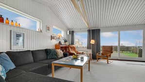 Un salon moderne avec un canapé gris, une table basse en bois et un fauteuil en cuir. On y trouve une bibliothèque avec des objets décoratifs et une grande fenêtre donnant sur l’extérieur.