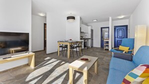 Ein modernes Wohnzimmer mit Flachbildfernseher, einem Esstisch mit Stühlen, einer Couch mit farbigem Kissen und einer Küchenzeile im Hintergrund.