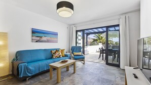 Ein modernes Wohnzimmer mit einem blauen Sofa, einem Couchtisch und einem Fernseher. Es gibt einen Balkon mit Gartenmöbeln und Meerblick.