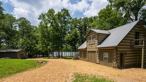 Une cabane rustique en rondins de bois, avec une allée de gravier, une grange en bois et un paysage boisé en arrière-plan.