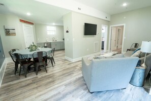 A modern living room with a grey sofa, a dining area with a table and chairs, a kitchen with a TV, and a hallway leading to another room. - Lake living at its best! (Cedar Bluff)
