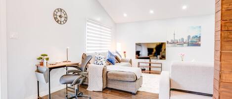 A modern living room with a sofa, a desk, and a chair. There is a clock on the wall and a window with blinds.