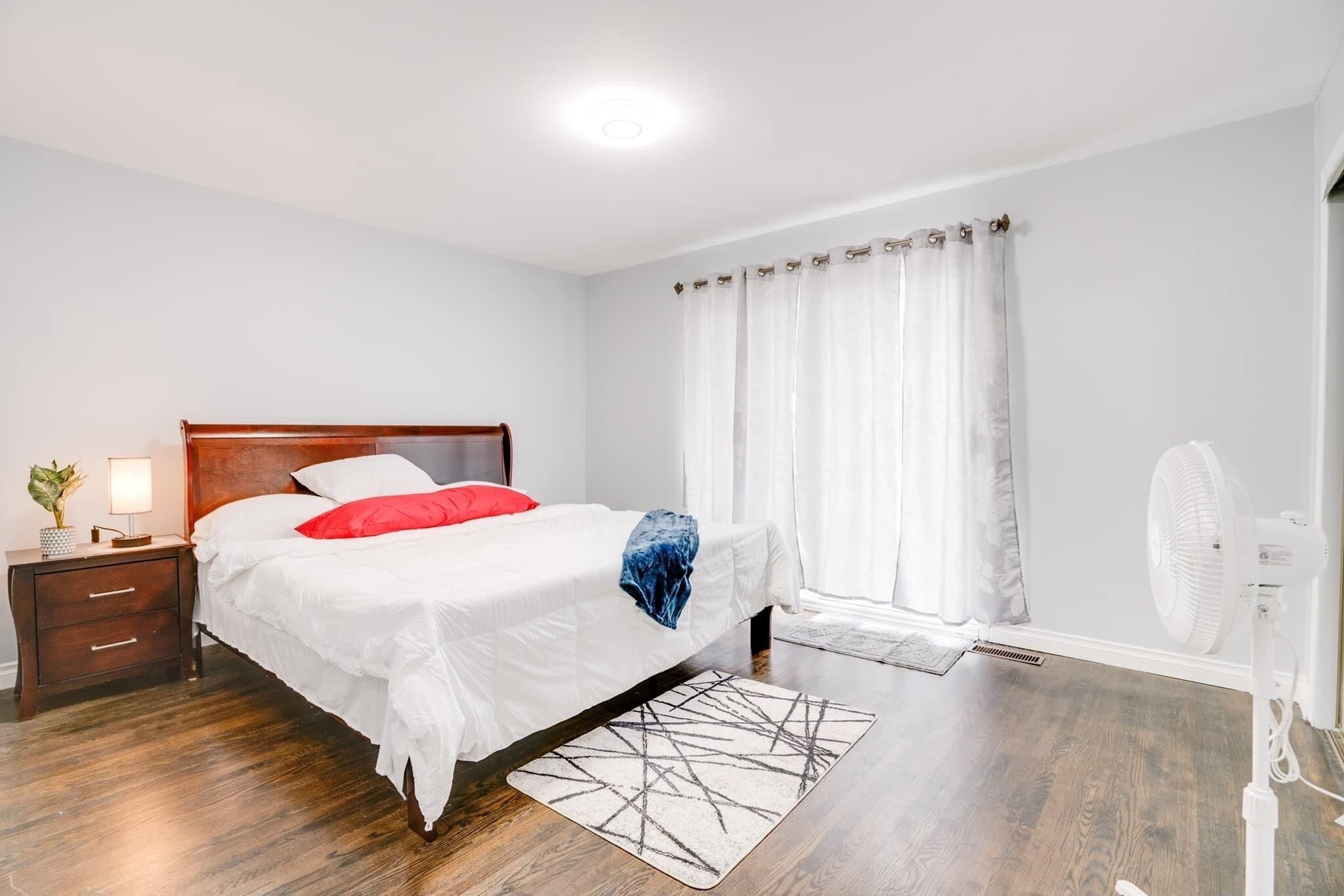 Ein Schlafzimmer mit einem hölzernen Bett, weißer Bettwäsche, einem roten Kopfkissen, einem Nachttisch mit Lampe, einer Stehlampe und einem Fenster mit weißen Vorhängen.