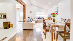 A spacious living area with a sofa, dining table, and chairs. There is a kitchen in the background, a chandelier, and a window with blinds. - Furnished Family Home ★ KING BED ★ Parking ★ BBQ ★ Backyard ★ Discounted Price (Pickering)