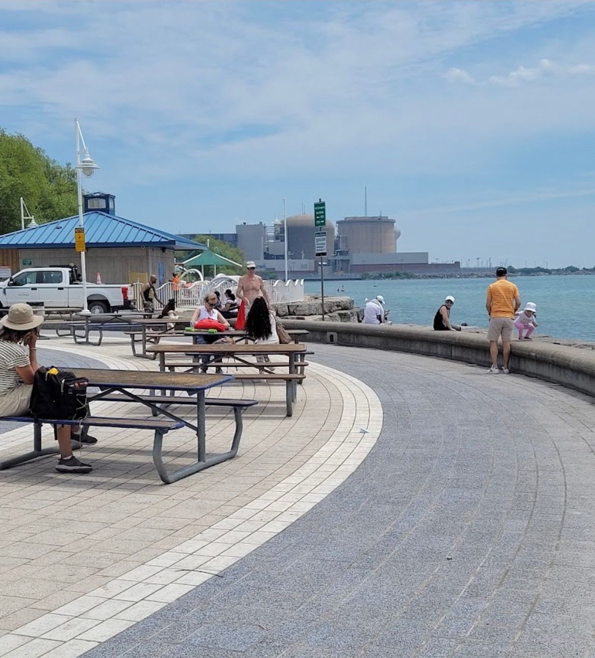 A waterfront promenade with benches and picnic tables, a clear sky, and a distant industrial skyline.