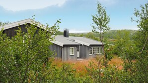 A rustic cabin with a sloped roof, surrounded by dense greenery and a hilly landscape in the background. - Awesome home in Ustaoset with WiFi (Ustaoset)