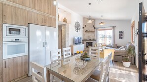 A modern kitchen with a built-in oven, microwave, and refrigerator. There is a dining area with a glass table and white chairs. A living room with a sofa and a fireplace is visible in the background.