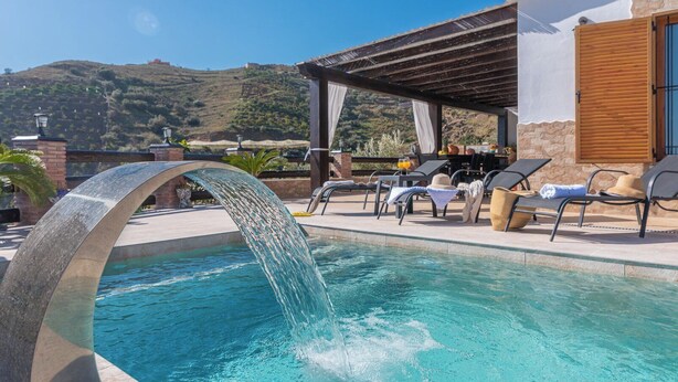 A pool with a water feature, lounge chairs, and a view of a hillside.