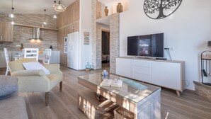 A modern living room with a glass coffee table, a sofa, a TV on a cabinet, and a kitchen in the background.