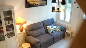 A living room with a grey sofa, two wooden stools, a glass cabinet with dishes, a table lamp, and a framed picture on the wall. - Bay view house in the heart of the Abbaye district (Saint-Valery-sur-Somme)