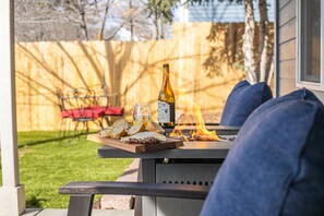 An outdoor patio with a table set for a meal, including bread, cheese, and a bottle of wine, with a fire in the background. - Luxury Downtown Cottage w/ Hot Tub, Private Yard, Fireplace & Fire Pit (Colorado Springs)
