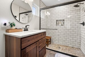 A modern bathroom with a wooden vanity, a round mirror, and a glass shower enclosure. - Luxury Downtown Cottage w/ Hot Tub, Private Yard, Fireplace & Fire Pit (Colorado Springs)