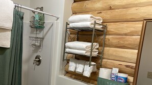 A bathroom with a wooden ceiling, a white toilet, a shower curtain, a towel rack with folded towels, and a shelf with toiletries. - Studio Style Cabin - In the heart of all things outdoors (Dillon)
