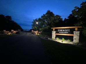 Een omheind toegangspunt naar Stone Gate Vacation Cabins, met een naambord, een verharde oprit en omliggend groen.