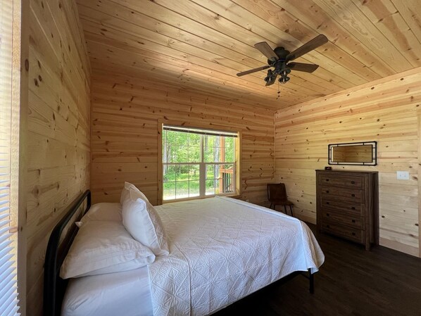 Un dormitorio con techo de madera, una cama con ropa de cama blanca, un aparador y una ventana con vistas a zonas verdes.