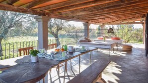 Une terrasse extérieure avec une table en bois dressée pour le repas, un banc et un lit agrémenté de coussins.
