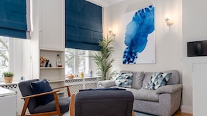 Ein modernes Wohnzimmer mit einem grauen Sofa, zwei Sesseln, einem blauen abstrakten Gemälde und einer Topfpflanze.