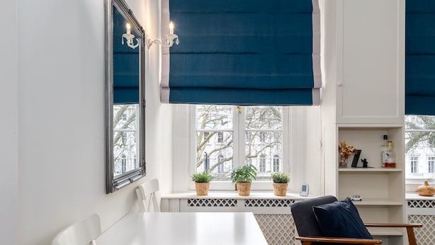Ein Zimmer mit einem weißen Tisch, einem dunklen Stuhl, einem Fenster mit blauen Jalousien und einem Regal mit Dekoration.