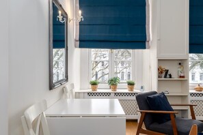 A room with a white table, a dark chair, a window with blue blinds, and a shelf with decorative items.