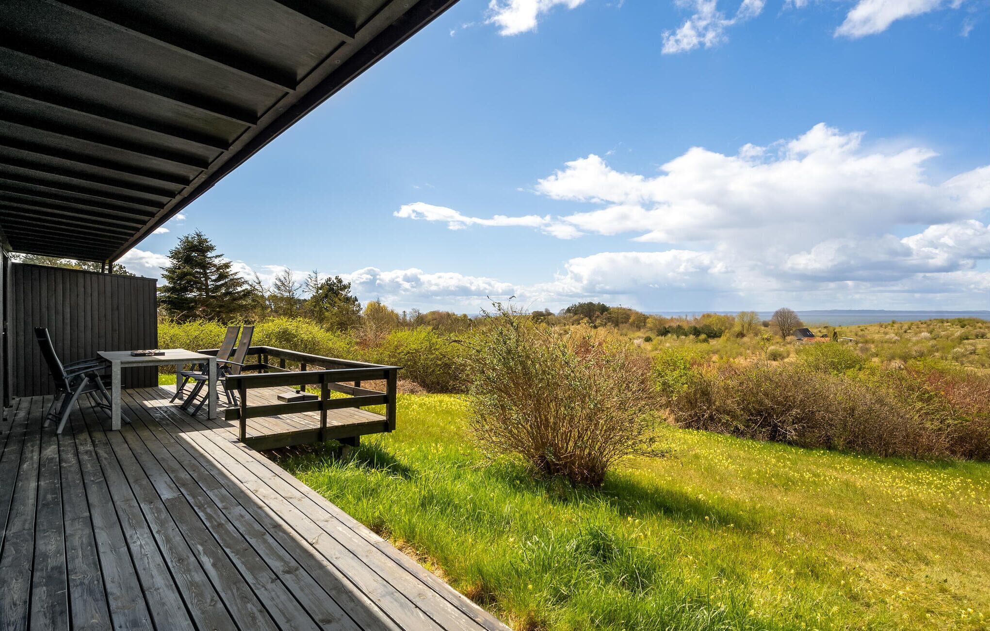 A wooden deck with a table and chairs, overlooking a grassy landscape with bushes and a distant view of the horizon.
