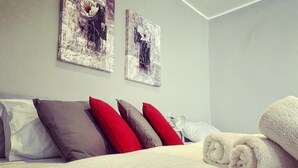 Ein ordentlich bezogenes Bett mit weißen und grauen Textilien, roten und grauen Kissen und aufgerollten weißen Handtüchern. Über dem Bett hängen zwei abstrakte Gemälde.