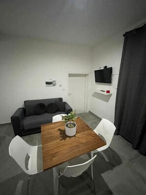 Un petit espace de vie moderne, avec une table en bois, des chaises blanches, un canapé gris, une plante en pot, un téléviseur posé sur une étagère et une porte.