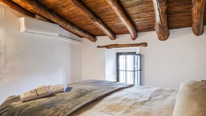 Een slaapkamer met een houten plafond, een bed met een geborduurde spreideken, een raam met tralies en een deur.