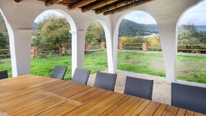 Een overdekt terras met een houten tafel en stoelen, omringd door witte zuilen, met uitzicht op een groen landschap en heuvels.