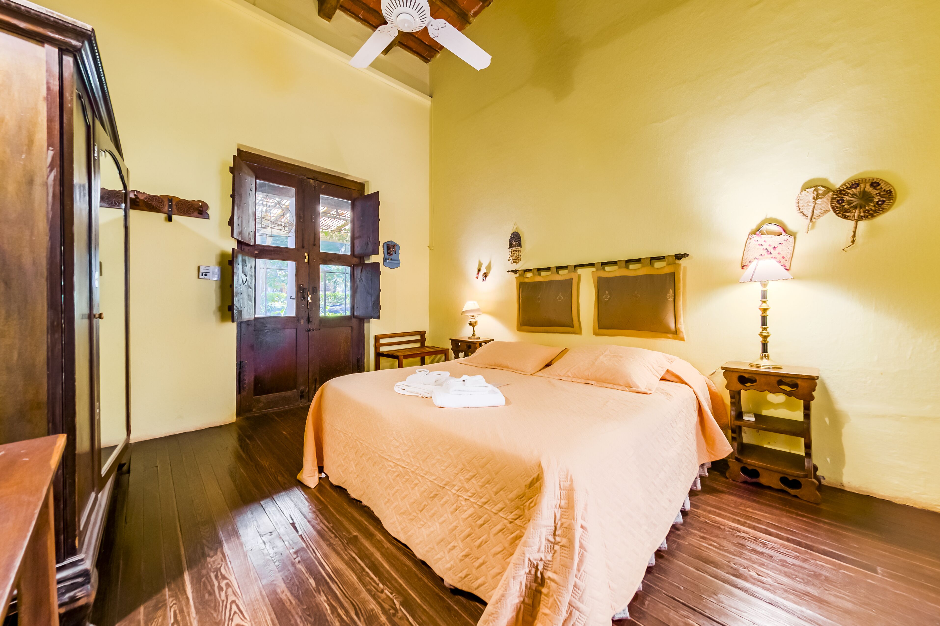 Une chambre avec un lit en bois, une armoire en bois, un ventilateur de plafond et une petite table avec une lampe.