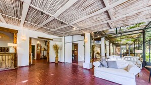 Eine geräumige Lobby mit rotem Backsteinboden, weißen Wänden und einer Decke mit freiliegenden Holzbalken. Im Sitzbereich befinden sich eine weiße Couch und Korbmöbel.