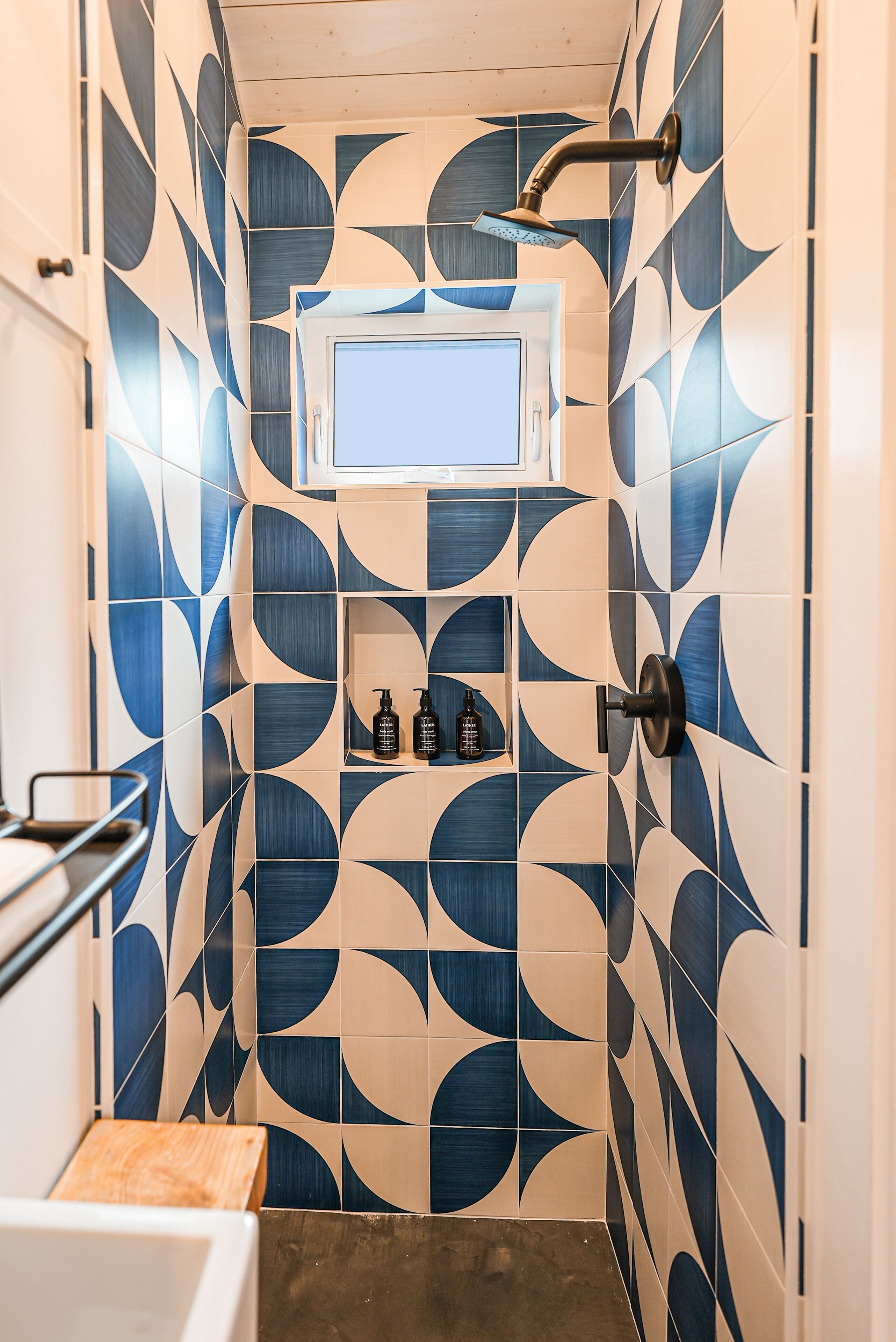 Un baño con un patrón geométrico de azulejos, un cabezal de ducha, una ventana pequeña y un estante con artículos de aseo.