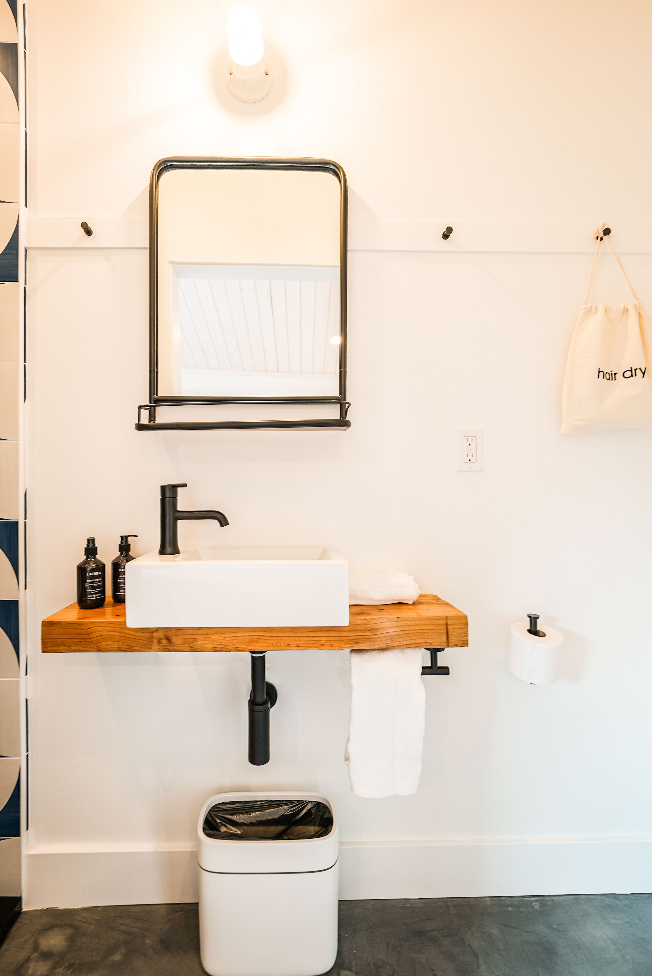 A bathroom with a white sink, black faucet, and a wooden shelf. There is a trash can and a towel hanging on the wall.