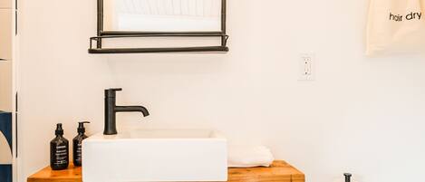 A bathroom with a white sink, black faucet, and a wooden shelf. There is a trash can and a towel hanging on the wall.