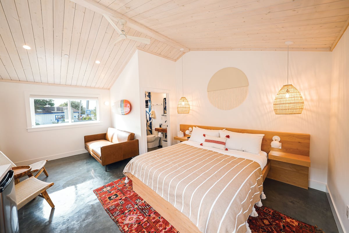 Une chambre avec un grand lit, une tête de lit en bois, une table de chevet, une chaise, un bureau, une fenêtre et un ventilateur de plafond.
