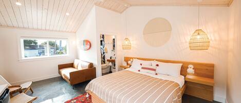 A bedroom with a large bed, a wooden headboard, a bedside table, a chair, a desk, a window, and a ceiling fan.