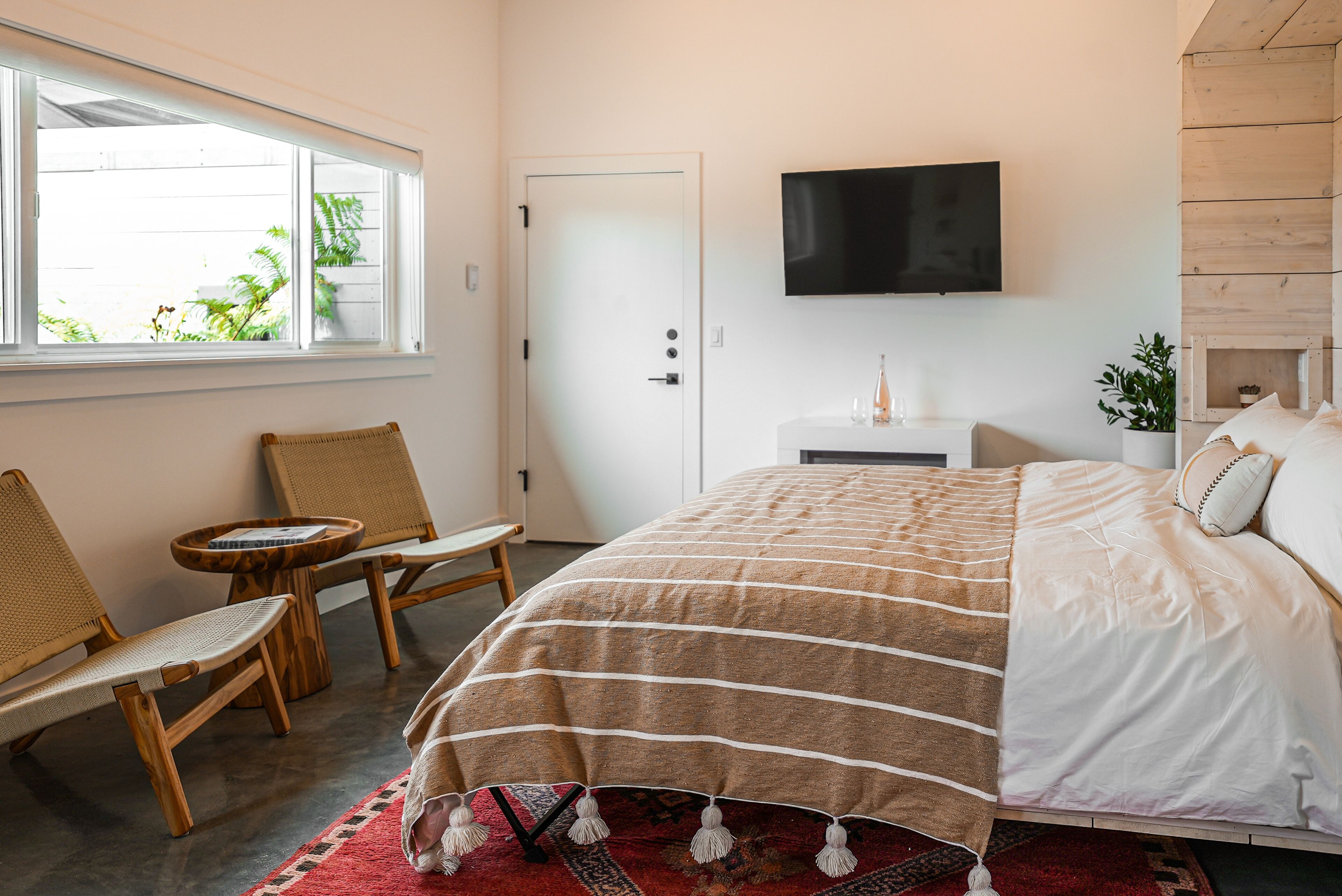 Un dormitorio con cama, televisor, ventana con vista a follaje, cabecera de madera y pequeña mesa con sillas.