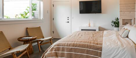 Un dormitorio con cama, televisor, ventana con vista a follaje, cabecera de madera y pequeña mesa con sillas.