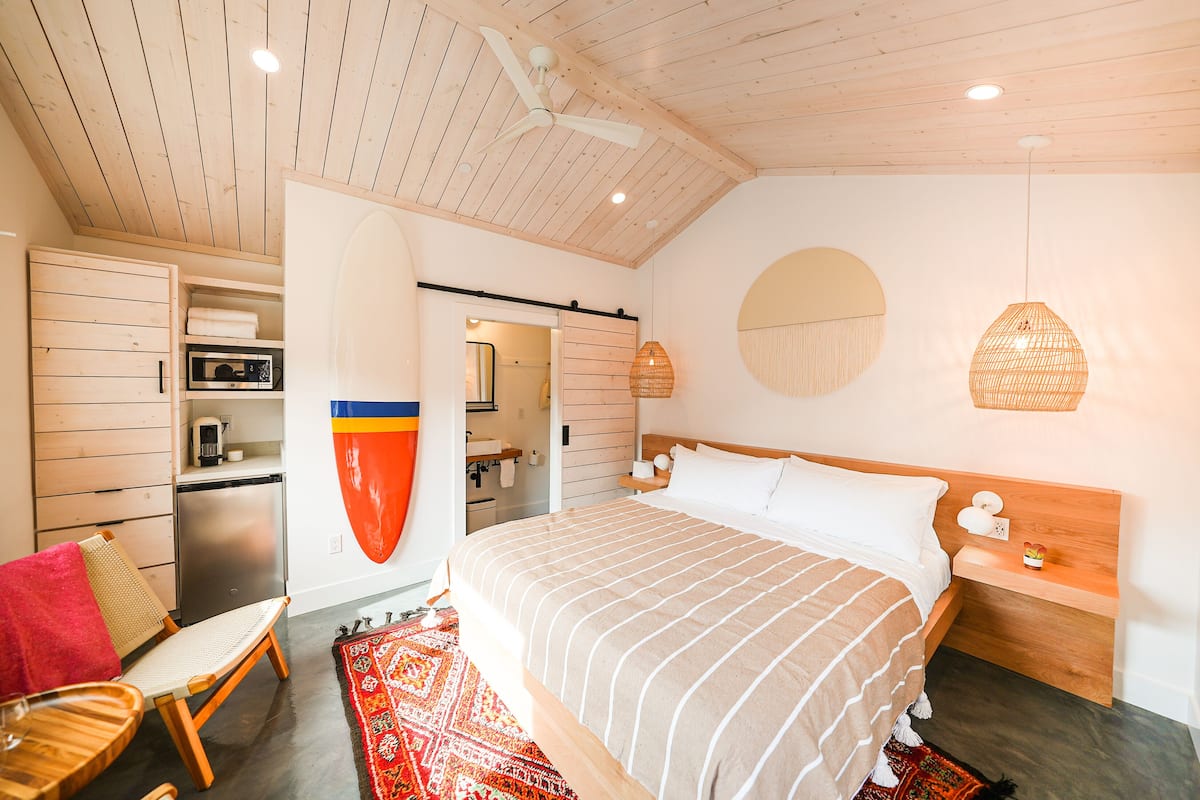 Une chambre moderne avec un plafond en bois, un lit recouvert d’une courtepointe à rayures, une petite table de chevet, une chaise, un four à micro-ondes et une serviette rouge.