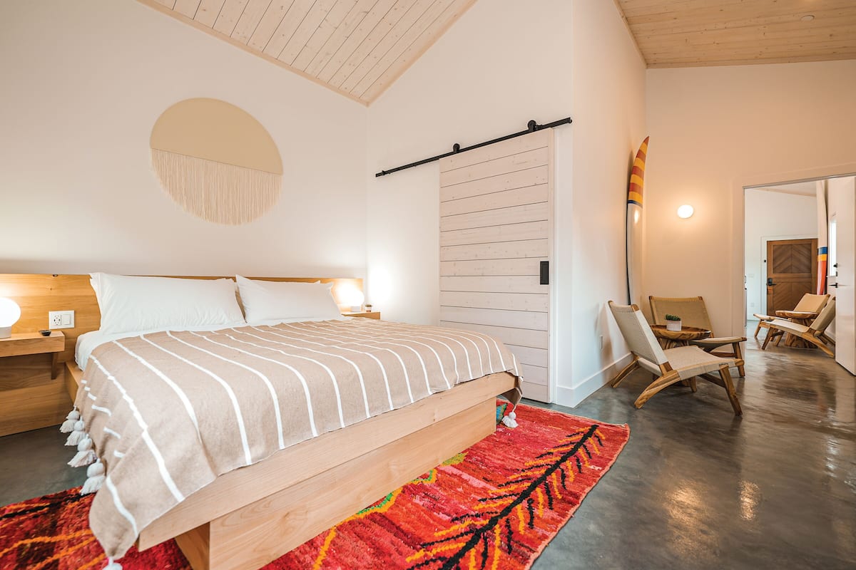 Une chambre à coucher comprenant un lit, des tables de chevet, une tête de lit, un luminaire suspendu, une porte coulissante et une chaise dans la pièce adjacente.